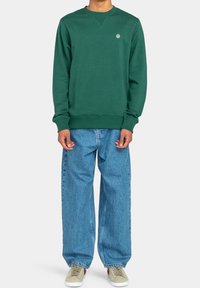 Persona che indossa un felpa girocollo verde scuro, jeans chiari larghi e sneakers beige con dettagli rossi, in piedi davanti a uno sfondo neutro.