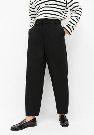 Persoon die zwarte broek met hoge taille en wijde pijpen draagt, gecombineerd met een zwart-wit gestreepte top met lange mouwen en zwarte loafers. Handen in de zakken.