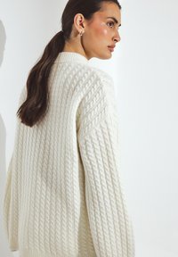 Woman with dark ponytail wearing a cream cable-knit sweater, looking over her shoulder against a plain white background.