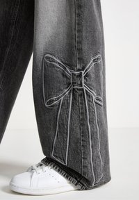 Szare spodnie dżinsowe z wyraźnym, wyszywanym wzorem kokardy po boku. W zestawieniu z białymi sneakersami, prezentując swobodny krój i detale przy nogawkach.