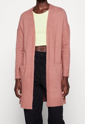 Personne portant un cardigan ouvert rose doux, un top court jaune clair et un pantalon noir taille haute sur un fond uni.
