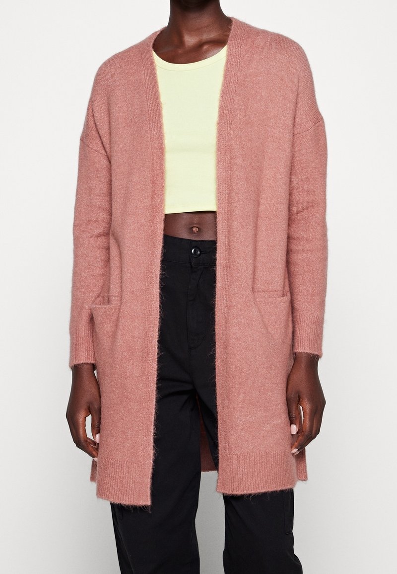 Personne portant un cardigan ouvert rose doux, un top court jaune clair et un pantalon noir taille haute sur un fond uni.