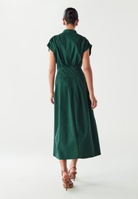 Žalia iki kelių ilgio suknelė su trumpomis rankovėmis, susiaurinta juosmeniu ir lygia tekstūra. Turi paprastą dizainą su užtrauktuku gale.