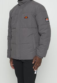 Grå polstret jakke med høj krave, prydet med et broderet logo på brystet og en sidelomme. Stoffet har en glat tekstur.