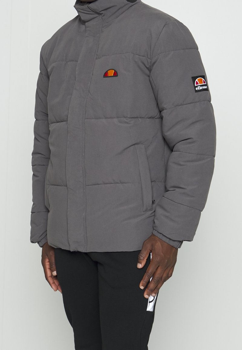 Grå polstret jakke med høj krave, prydet med et broderet logo på brystet og en sidelomme. Stoffet har en glat tekstur.
