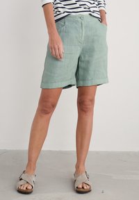 Světlé zelené lněné šortky s ohrnutým lemem, mají přední kapsy a zapínání na knoflík. Doplněno světlými šedými sandály.