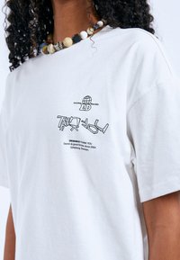 Bílá bavlněná trička s krátkým rukávem. Vybaveno grafickým potiskem černým textem a designem globu na hrudi, který ukazuje uvolněný styl.