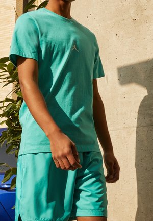 Mand iført teal Jordan T-shirt og matchende shorts, stående udendørs nær en betonvæg med skygge og en grøn plante synlig.