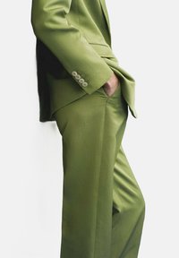 Olive green suited outfit with a fitted blazer and tailored trousers. Smooth fabric, single button detail on sleeves, and side pockets.