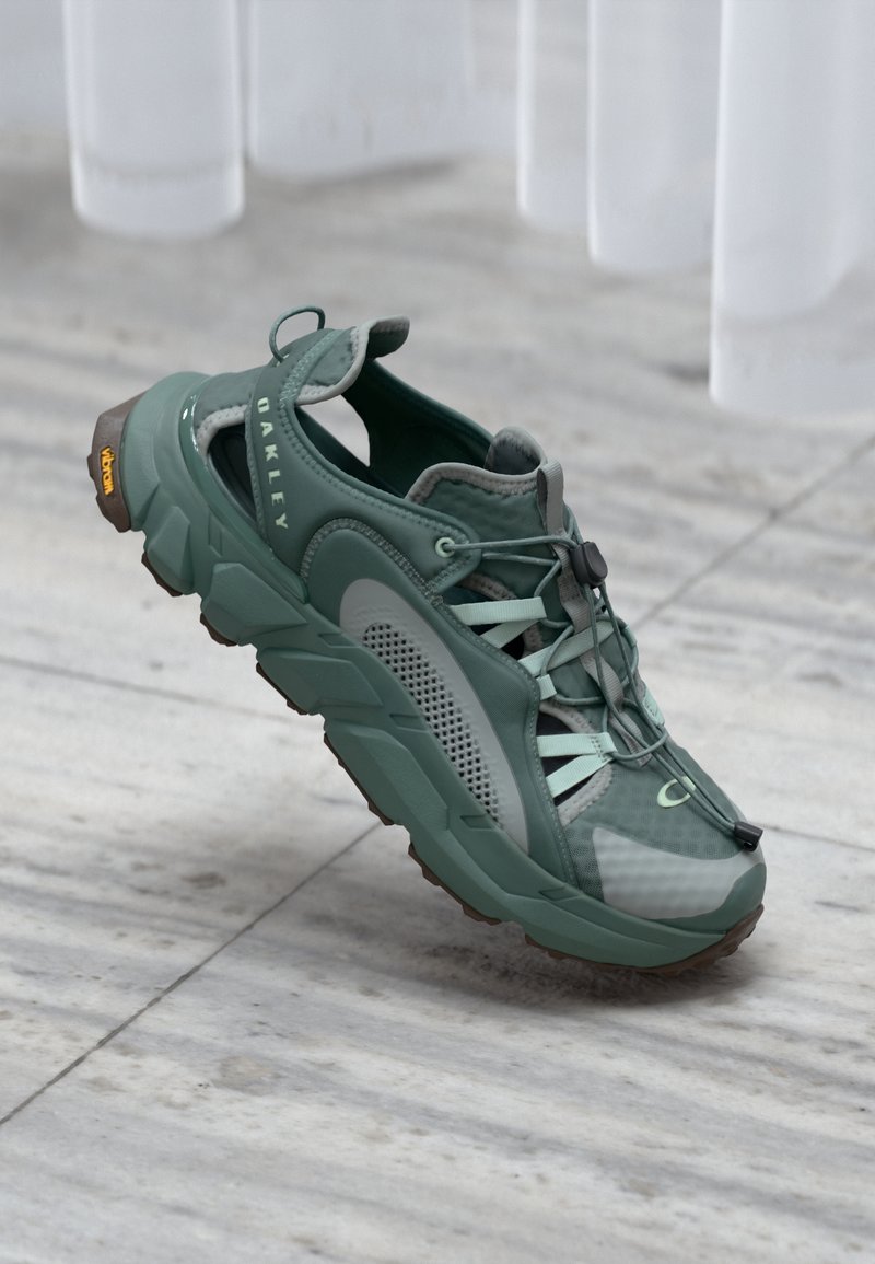 Scarpa sportiva verde Oakley con pannelli in mesh, sistema di allacciatura rapida e suola Vibram su uno sfondo in marmo.