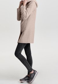 Beige hooded coat with a tailored fit, paired with dark skinny jeans and gray sneakers featuring textured accents and white soles.