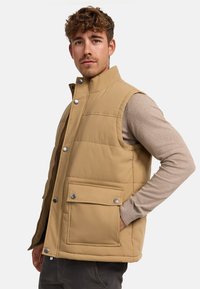 Gilet matelassé beige avec un col montant, des boutons-pression et une poche frontale. Fabriqué en tissu lisse avec une doublure intérieure confortable.