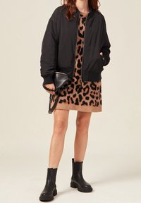 Femme portant une veste bomber noire sur une robe à imprimé léopard, tenant un sac à main noir et portant des bottines noires.