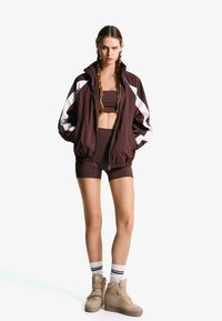 Jachetă bomber maro, cu fermoar, cu dungi albe, asociată cu un top scurt maro și pantaloni scurți. Sneakers bej cu vârf înalt și curea pentru gleznă.