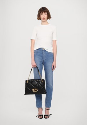 Borsa a tracolla nera trapuntata con logo dorato, tenuta da una persona in camicia bianca aderente e jeans blu. I sandali presentano un accento a fiocco.