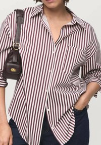 Camisa de botones a rayas en burdeos y blanca, con un bolsillo en el pecho, corte relajado, mangas largas y puños enrollados. Combinada con un bolso marrón.