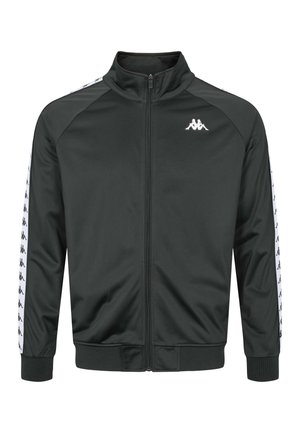 Man draagt een zwarte trainingsjas met rits en bijpassende broek met witte logostrepen over een wit T-shirt, staand tegen een effen achtergrond.