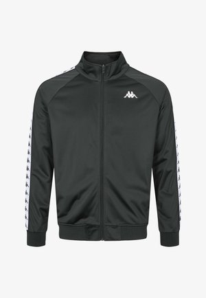 Mężczyzna noszący czarną sportową kurtkę na zamek błyskawiczny oraz pasujące spodnie z białymi paskami z logo na białej koszulce, stojący na jednolitym tle.