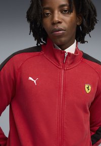 Rød zip-up jakke med sorte skulder accenter, et hvidt Puma-logo og et gult Ferrari-emblem. Blødt tekstureret stof, atletisk design.