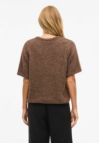 Kort brun strikkegenser med korte ermer, rund hals og avslappet passform. Teksturen virker myk og lodden. Sorte bukser synlige nedenfor.