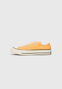 Oranžid kootsahased sneakerid valgete paeltega, must kummist varbakiil ja kahekordne must riba kreemilisel tallal. Minimalistlik disain, madal profiil.