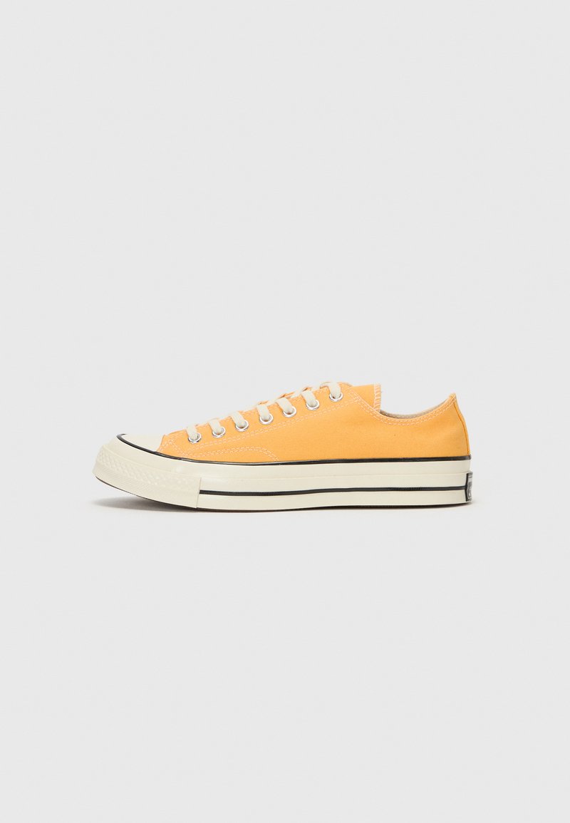 Orange canvas sneaker with white laces, black rubber toe cap, and dual black stripes on the creamy sole. Minimalist design, low profile.