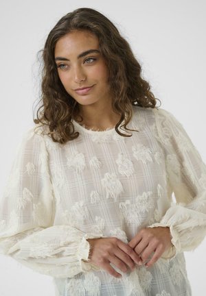 Camicia bianca con texture e finitura trasparente, caratterizzata da un motivo a quadri, maniche a puff e polsini arricciati, decorata con tocchi floreali sparsi.