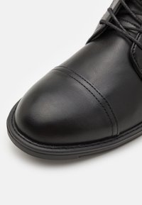 Gros plan de l'avant d'une chaussure habillée en cuir noir avec bout renforcé cousu et lacets noirs sur fond blanc.
