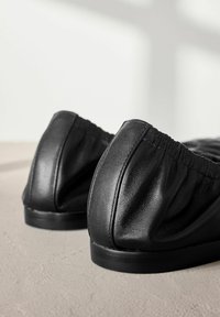 Schwarze Leder-Ballettschuhe mit gerafftem Absatz, glatter Textur und flacher Gummisohle, auf einer neutralen Oberfläche platziert.