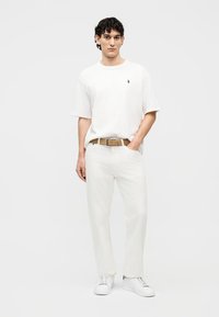 Joven con cabello rizado y oscuro, vestido con camiseta blanca, pantalones blancos, cinturón beige y zapatillas blancas, de pie con una mano en el bolsillo.