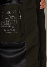 Waterdichte shell-stof jas in donkergroen, met een ritszak, ribgebreide manchetten en een binnenlabel met ontwerp-instructies en logo.