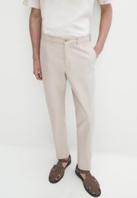 Ljusbeige byxor med en slät yta, rak passform, framfickor och ett matchande bälte. Kombinerade med bruna, remmade sandaler.