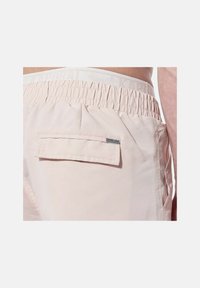 Shorts roses clairs avec une taille élastique, présentant une poche arrière avec un rabat et une étiquette de logo. Texture lisse avec un design minimaliste.