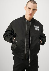 Čierna bomber bunda z hladkej látky, s predným zipsom, bočnými vreckami a strieborným logom na hrudi. Rúbkované manžety a lem.