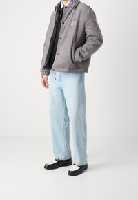 Grå kordfløjl jakke med ribbet tekstur, knaplukning og logo detaljer, parret med lyseblå jeans og sorte loafers med hvide accenter.