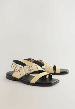 Žluté kožené sandály se stříbrnými detaily, mají křížové pásky, kruhové výřezy a plochou černou podrážku.