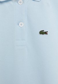 Světle modré polo tričko z texturované látky s tříknoflíkovým zapínáním a malým zeleným krokodýlím logem na levé straně hrudi.