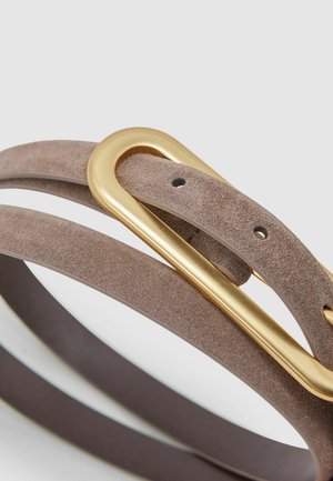 Šviesiai rudos spalvos zomšiniai diržas su lygiu, apvaliu auksiniu sagtimi. Turi keletą skylių reguliavimui ir yra plokščias, lieknas dizainas.