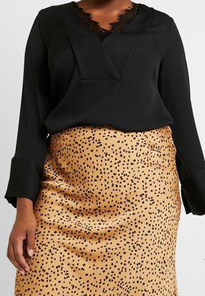 Persoon die een zwarte blouse met lange mouwen draagt met een kanten halsdetail en een beige satijnen rok met een zwart gevlekt patroon.
