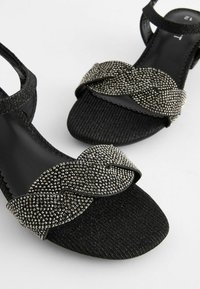 Sorte sandaler med en struktureret rem, der har et krydset design prydet med sølv rhinesten. Flad sål med et slip-on design.