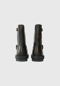 Un par de botas de cuero marrón oscuro hasta el tobillo con hebillas laterales, vistas desde la parte posterior, sobre un fondo claro y sencillo.
