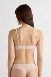 Vrouw met lang bruin haar draagt een beige kanten lingerieset, gezien van achteren tegen een effen witte achtergrond.