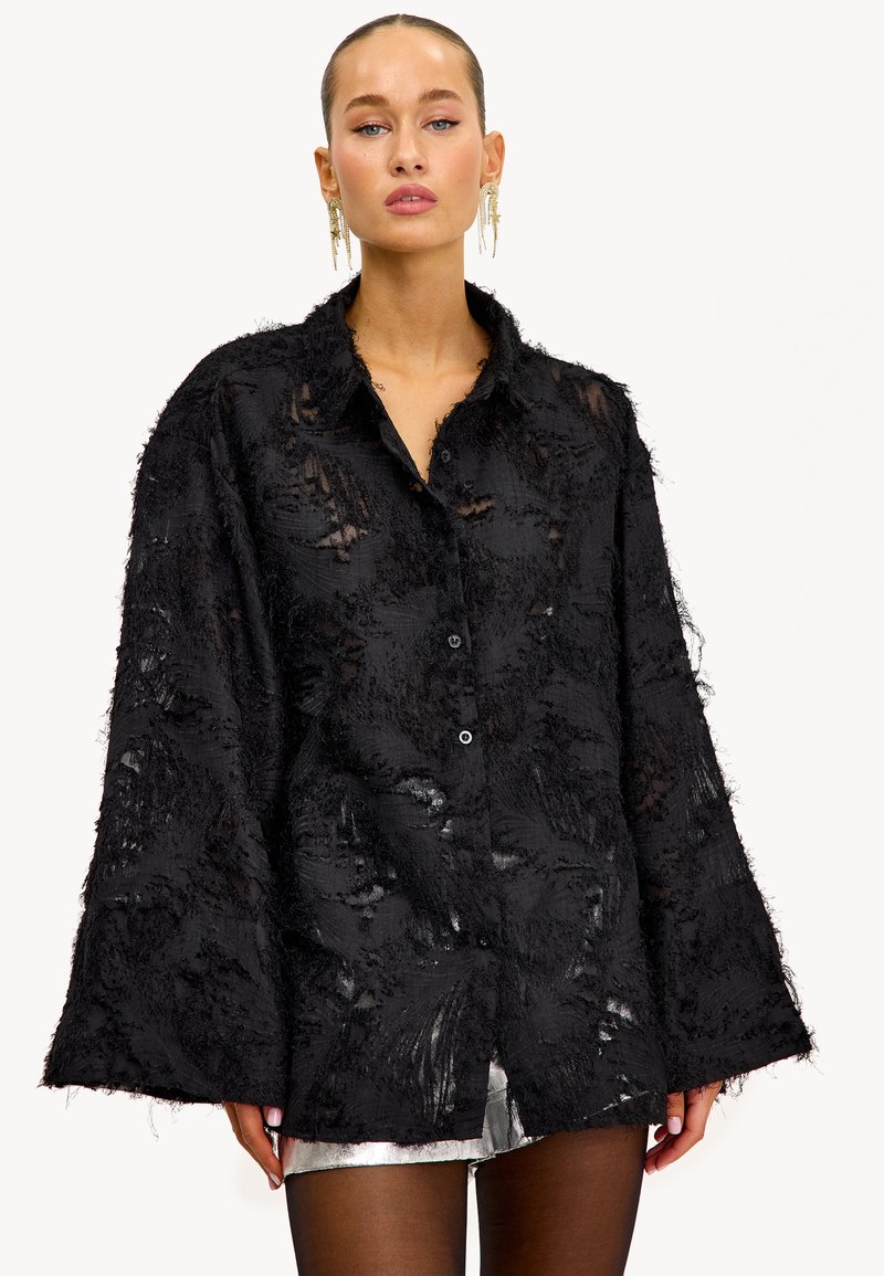 Zwarte oversized shirt met textuurdetails en rafelige randen, met een knoopsluiting aan de voorkant en een relaxte pasvorm. Gecombineerd met zilveren shorts.