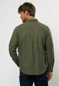 Olīvzaļš garām piedurknēm krekls ar klasisku apkakli, pogu aprocēm un muguras šuvi. Mīksts audums ar gludu tekstūru.