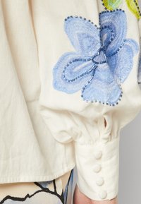 Witte blouse met blauwe bloemenborduring op de mouw; heeft een gathered pofmouw, een getailleerde manchette met vier stoffen knopen, zachte textuur.