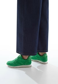 Sneaker in suede verde con suola bianca, caratterizzate da cuciture intricate e un design elegante, abbinate a pantaloni in denim scuro larghi.