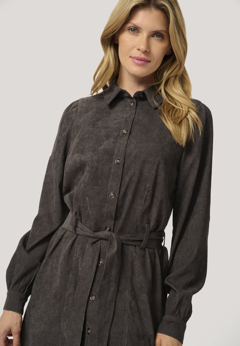 Femme blonde portant une robe boutonnée gris foncé avec de longues manches bouffantes et une ceinture en tissu nouée à la taille.