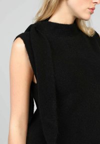 Pull noir sans manches avec un col montant, fabriqué en tissu texturé doux. Présente un détail drapé sur une épaule et des emmanchures ouvertes.