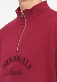 Hanorac bordeaux cu guler înalt, fermoar argintiu și text brodat "ORIGINALS Studio" într-o nuanță ușor mai închisă. Textură moale.
