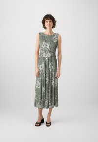 Robe maxi florale en vert atténué avec un détail de corsage torsadé, jupe évasée et sandales à talons blocs noires. Tissu doux avec une texture légère.